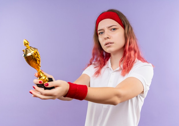 Free Photo young sporty woman in headband holding trophy with serious expression standing over purple wall