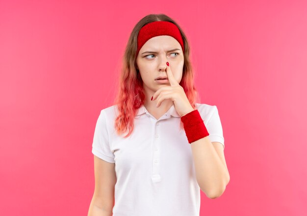 Young sporty woman in headband looking aside touching her nose with pensive expression on face standing over pink wall