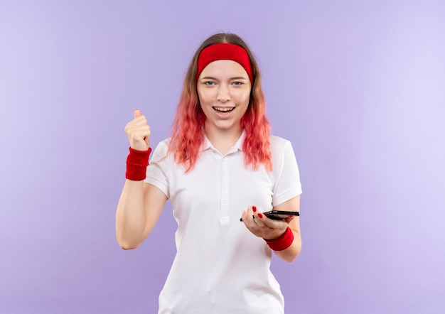 Free Photo young sporty woman holding smartphone clenching fist smiling cheerfully standing over purple wall