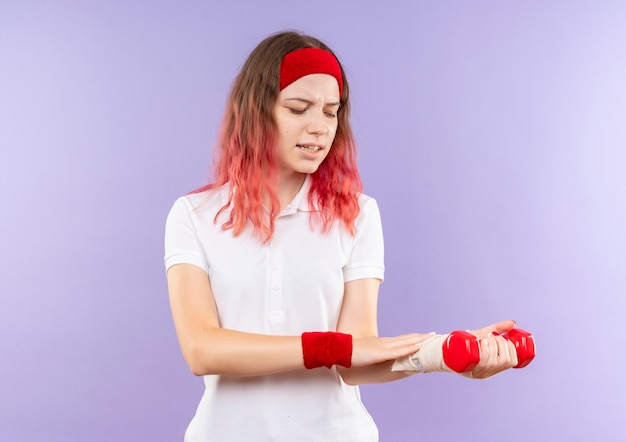 Free photo young sporty woman holding two dumbbells doing exercises looking at her bandaged wrist feeling pain standing over purple wall