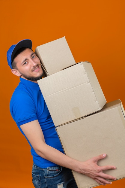 Free Photo young over strained delivery man wearing blue polo shirt and cap standing with stack of boxes looking tired over isolated orange background