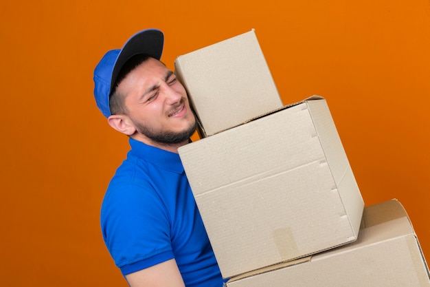 Young over strained delivery man wearing blue polo shirt and cap standing with stack of boxes looking tired over isolated orange background