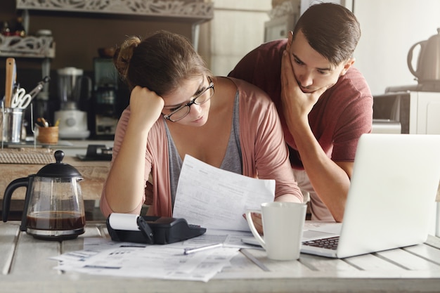 Free photo young stressed caucasian couple facing financials troubles, sitting at kitchen table with papers, calculator and laptop computer and reading document from bank, looking frustrated and unhappy