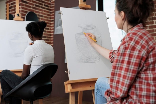 Young student drawing artistic vase on painting canvas working at illustration technique using graphic pencil during art lesson in creativity studio. Multiethnic team developing creative skill