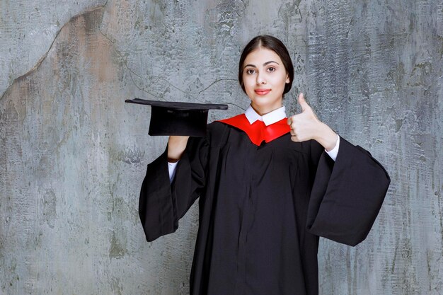 Young student in gown holding hat and giving thumbs up. High quality photo