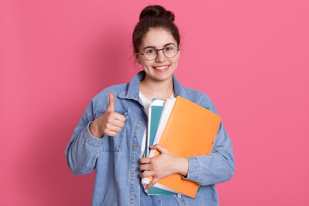 young student woman wearing denim jacket and eyeglasses, holding colorful folders and showing thumb up on pink