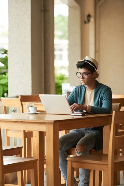 Young stylish Asian man sitting at table in cafe and working on laptop