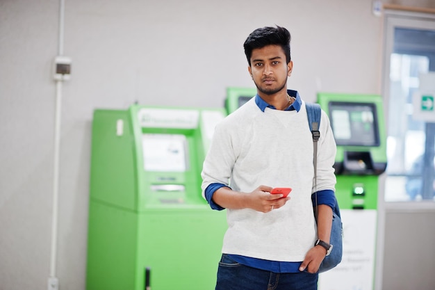 Free photo young stylish asian man with mobile phone and backpack against row of green atm