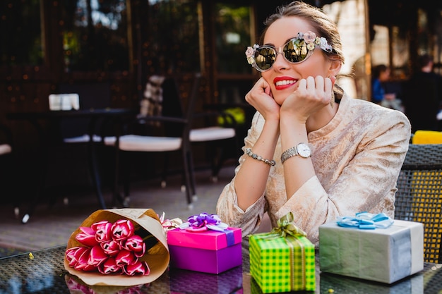 Free photo young stylish beautiful woman in fashion sunglasses sitting at cafe with presents