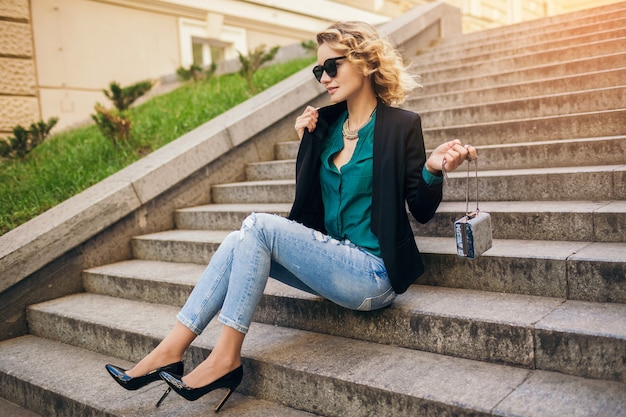 Free Photo young stylish beautiful woman sitting in street, wearing jeans, black jacket, green blouse, sunglasses