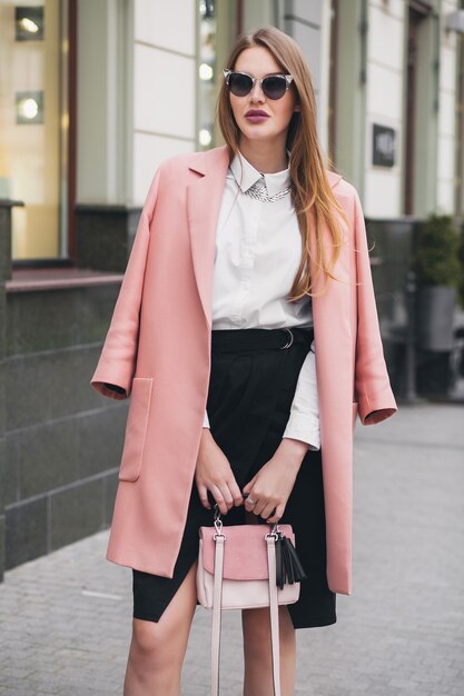 Young stylish beautiful woman walking in street, wearing pink coat, purse, sunglasses, white shirt, black skirt, fashion outfit, autumn trend, smiling happy, accessories