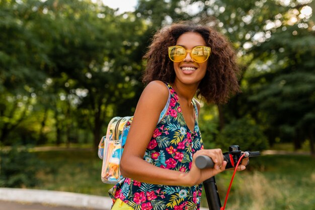 Young stylish black woman having fun in park riding on electric kick scooter in summer fashion style, colorful hipster outfit, wearing backpack and yellow sunglasses