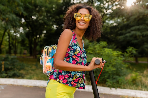 Young stylish black woman having fun in park riding on electric kick scooter in summer fashion style, colorful hipster outfit, wearing backpack and yellow sunglasses