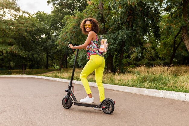 Young stylish black woman having fun in park riding on electric kick scooter in summer fashion style, colorful hipster outfit, wearing backpack and yellow trousers and sunglasses