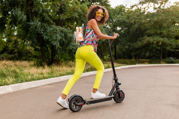 Young stylish black woman having fun in park riding on electric kick scooter in summer fashion style, colorful hipster outfit, wearing backpack and yellow trousers and sunglasses