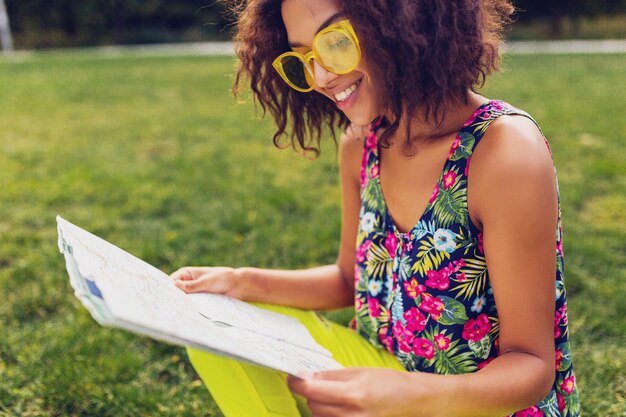 Young stylish black woman having fun in park summer fashion style, colorful hipster outfit, sitting on grass, traveler with a map