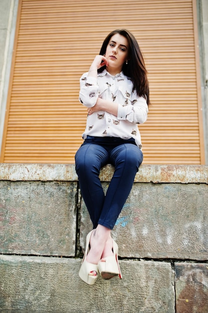 Young stylish brunette girl on shirt pants and high heels shoes sitting and posed background orange shutter Street fashion model concept