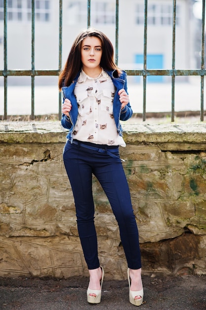 Young stylish brunette girl on shirt pants jeans jacket and high heels shoes posed background iron fence Street fashion model concept