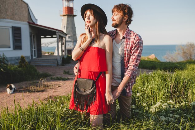 Young stylish couple in love in countryside, indie hipster bohemian style, weekend vacation, summer outfit, red dress, green grass, holding hands, smiling