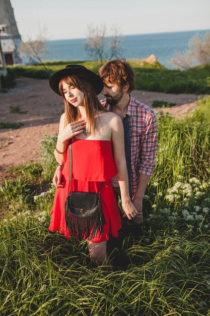 Young stylish couple in love in countryside, indie hipster bohemian style, weekend vacation, summer outfit, red dress, green grass, holding hands, smiling