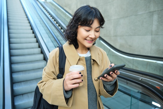 Free photo young stylish girl with cup of coffee drinks cappuccino to go goes down escalator and looks at mobil