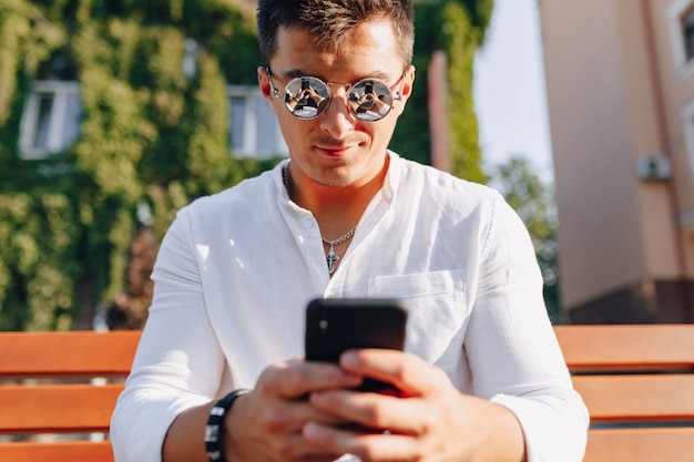 Young stylish guy in shirt with phone on bench on sunny warm day outdoors