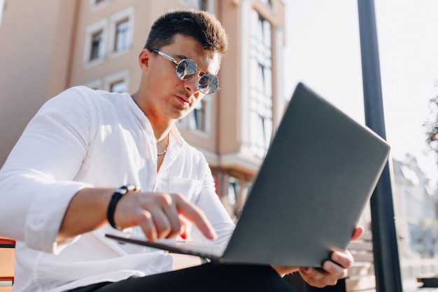 Young stylish guy in shirt with phone and notebook on bench on sunny warm day outdoors, freelance