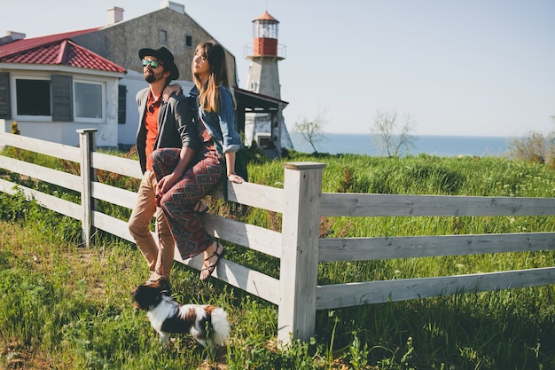 Young stylish hipster couple in love walking with dog in countryside, summer style boho fashion