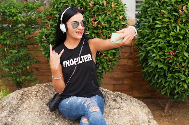 Free Photo young stylish hipster woman in black t-shirt, jeans, listening to music on headphones, having fun, posing, taking selfie photo on phone, showing peace sign, funny face expression