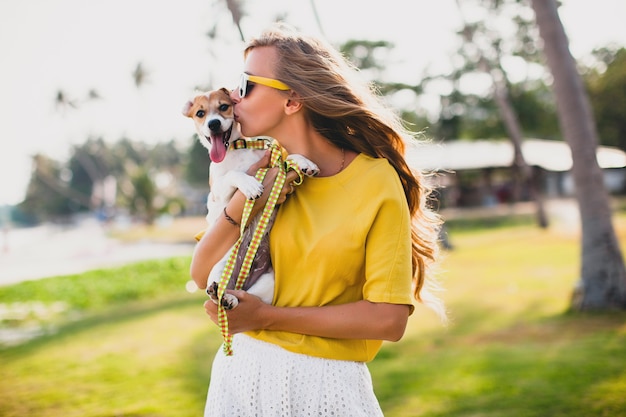 Young stylish hipster woman holding walking playing dog puppy jack russell, tropical park, smiling and have fun, vacation, sunglasses, cap, yellow shirt, beach sand