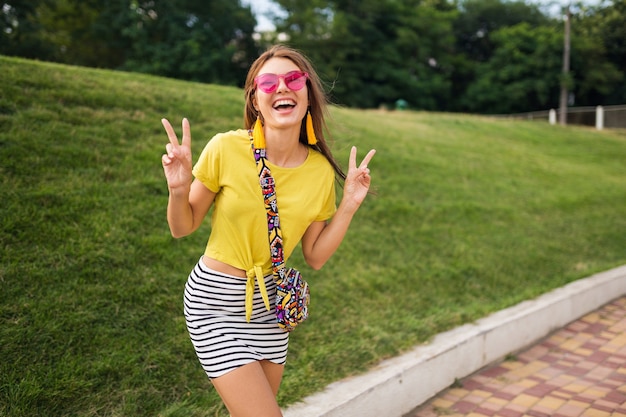 Young stylish laughing woman having fun in city park, smiling cheerful mood, positive, emotional, wearing yellow top, striped mini skirt, handbag, pink sunglasses, summer style fashion trend