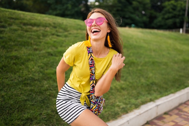 Young stylish laughing woman having fun in city park, smiling cheerful mood, wearing yellow top, striped mini skirt, pink sunglasses, summer style fashion trend