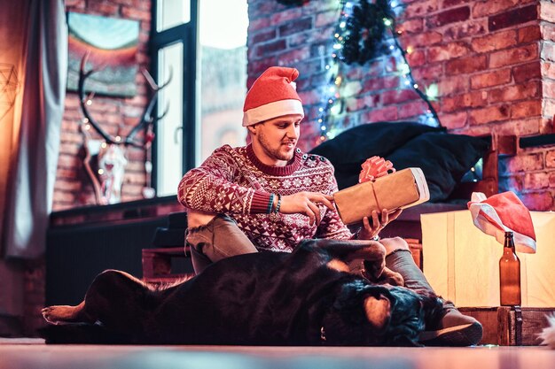 A young stylish man holding a gift box while sitting with his cute dog in a decorated living room at Christmas time.