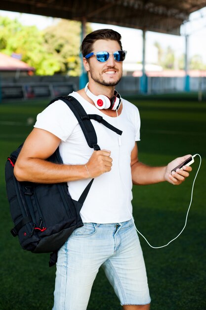 Young stylish man posing on a soccer field, listen music. Headphones on his shoulders, holding player in hand, sunglasses on the face