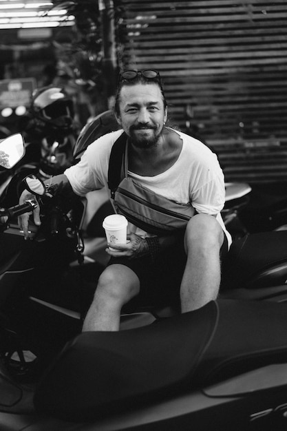 Free Photo a young stylish man with a beard drinks coffee from a disposable paper cup, sits on a motorbike on the street. lifestyle, tropical life. bali