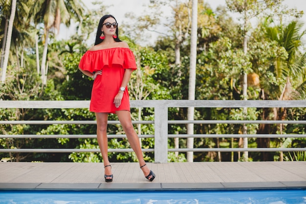 Free photo young stylish sexy woman in red summer dress standing on terrace in tropical hotel, palm trees background, long black hair, sunglasses, ethnic earrings, sunglasses, looking forward, high heel shoes