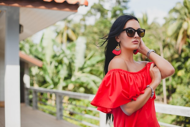 Young stylish sexy woman in red summer dress standing on terrace in tropical hotel, palm trees background, long black hair, sunglasses, ethnic earrings, sunglasses, smiling