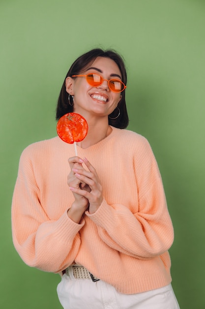 Young stylish woman in casual peach sweater and orange glasses isolated on green olive wall with orange lollipop positive smile copy space