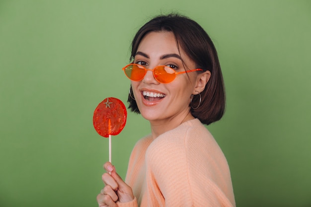 Free photo young stylish woman in casual peach sweater and orange glasses isolated on green olive wall with orange lollipop positive smile copy space