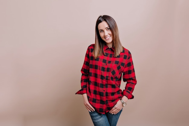 Free photo young stylish woman dressed checkered shirt and jeans poses on the isolated wall with charming smile and happy emotions