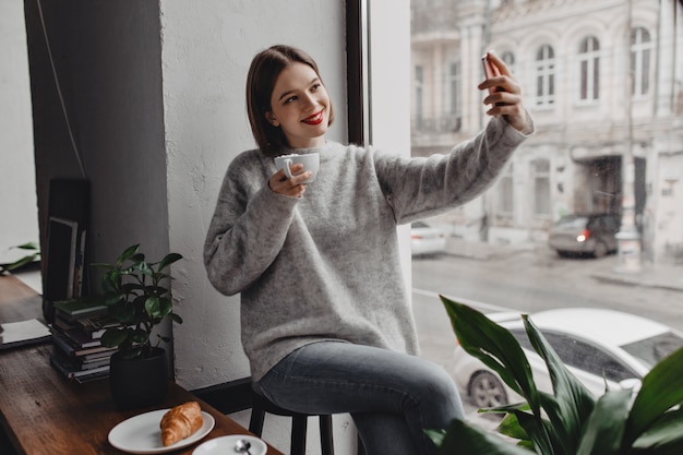 Free photo young stylish woman in gray sweater and jeans posing with cup of cappuccino and taking selfie by window.