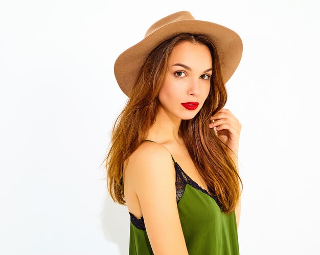 Young stylish woman model in casual summer green clothes and brown hat with red lips, posing near white wall