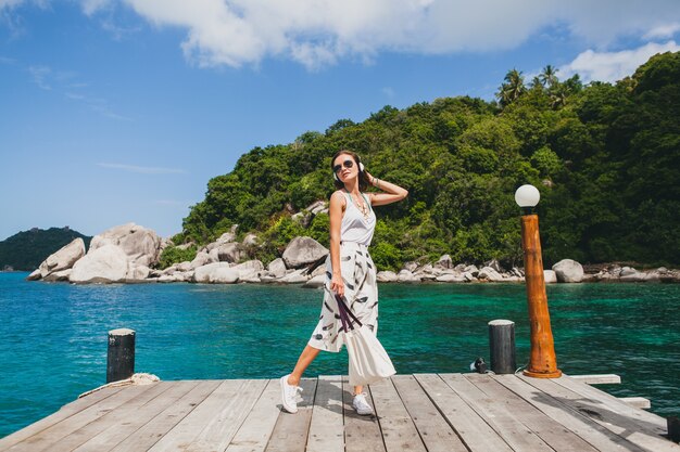 Young stylish woman standing on pier, walking, listening to music on headphones, summer apparel, white skirt, handbag, azure water, landscape background, tropical lagoon, vacation, traveling in asia