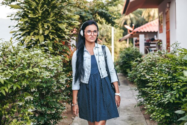 Young stylish woman walking with smartphone, listening to music on headphones, summer vacation