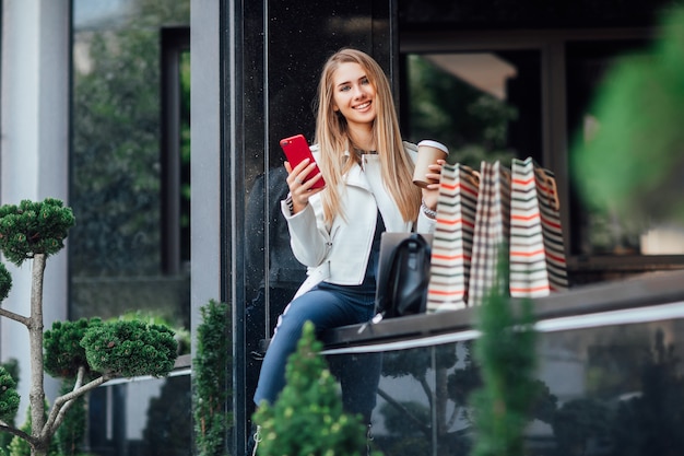 Free photo young, successful blonde fashionable woman with cup of coffee and phone, sit near store after her shopping time.