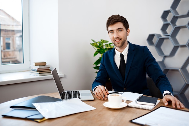 Young successful businessman sitting at workplace, office background.