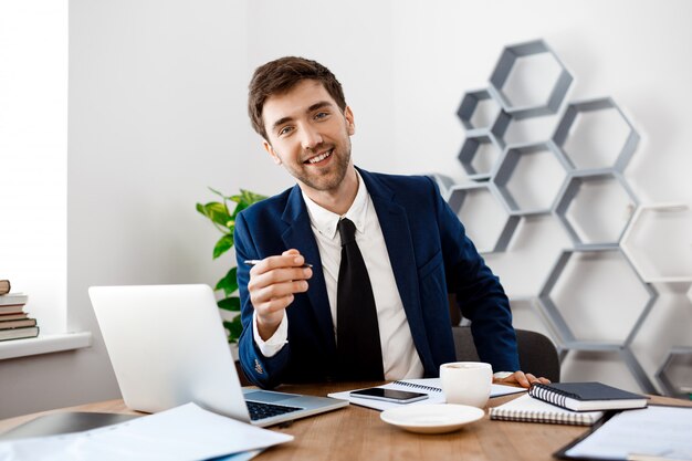 Young successful businessman sitting at workplace, office background.