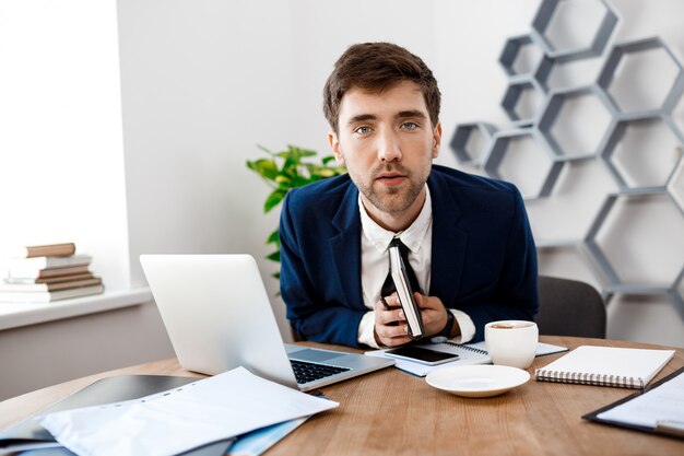 Young successful businessman sitting at workplace, office background.