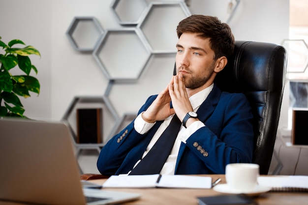 Young successful businessman sitting at workplace, office background.