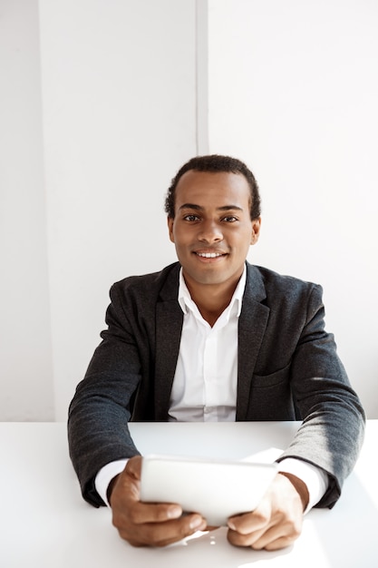 Young successful businessman smiling, holding tablet, sitting at workplace